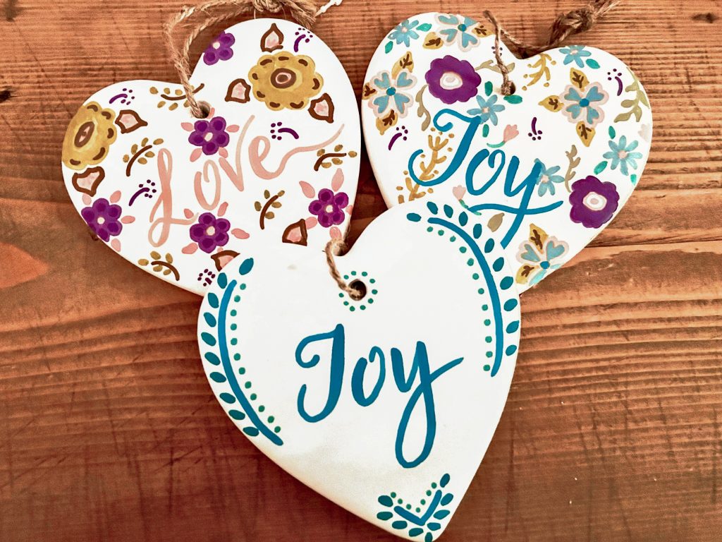 a trio of white hearts with joy and love written on them surrounded by painted flowers. they are laying on a wooden surface