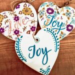 a trio of white hearts with joy and love written on them surrounded by painted flowers. they are laying on a wooden surface