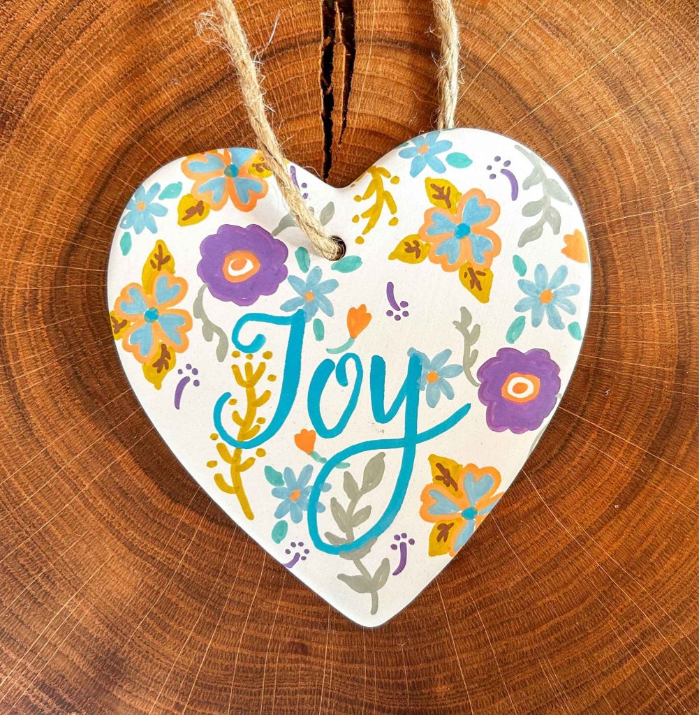 a white heart on a wooden background.  the heart is decorated with the word joy in blue and flowers around the word 