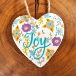 a white heart on a wooden background. the heart is decorated with the word joy in blue and flowers around the word
